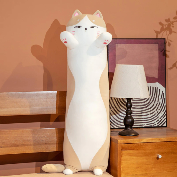 Sobre una cama con una mesilla de noche al lado, con una lámpara de mesilla blanca y un marco detrás, hay una almohada de gato blanco y marrón, de pie sobre sus pequeñas patas traseras