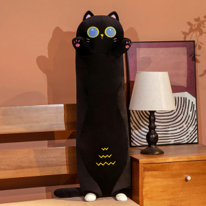 Sobre una cama junto a la que hay una mesilla de noche con una lámpara de mesilla blanca y un marco detrás, hay un cojín de felpa de gato negro, se sostiene sobre sus diminutas patas traseras