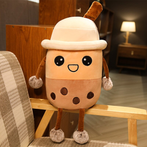 Peluche de té de burbujas con sombrero, es de color beige y marrón, y está sentada en el reposabrazos de madera de un sillón a cuadros beige, blanco y marrón en un piso con una lámpara encendida al fondo