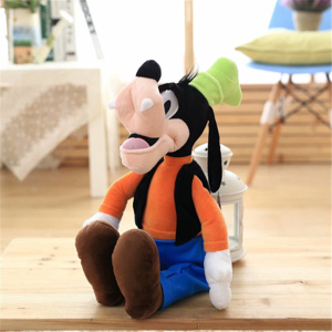 Peluche del personaje bobalicón de Disney sentado en un parquet, lleva pantalones azules y un top naranja
