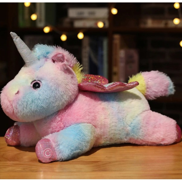 Sobre una mesa de madera un peluche de unicornio multicolor está tumbado boca abajo con estanterías y luces de fondo