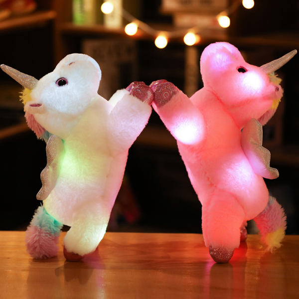 Sobre una mesa de madera, 2 peluches de unicornio, uno blanco y otro rosa están levantados sobre sus patas traseras y se sujetan por la delantera y sus cuerpos están iluminados